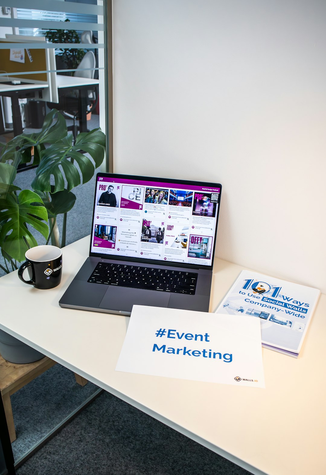 A desk with documents, a piece of paper with the hashtag #EventMarketing and a laptop showing a social media wall